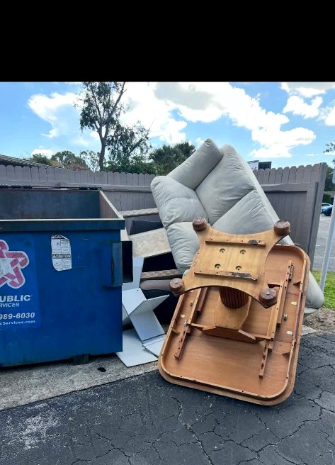 Bulk trash piled curbside next to a dumpster — table, furniture, and household debris